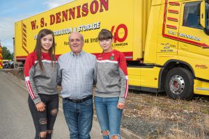 W.S. Dennison Transport Manager, Liam Taggart, with daughters Aoife and Caitlin standing in front of a yellow and red W.S. Dennison truck and trailer